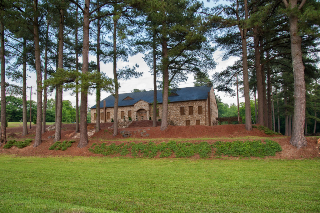 A rustic stone house surrounded by tall pine trees, with a lush green lawn in the foreground